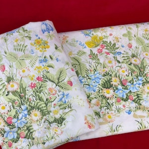 May include: White floral fabric featuring a vibrant pattern of wildflowers, including daisies, bluebells, and strawberries, with green foliage. The fabric is folded, showcasing the detailed design. The overall theme is nature and botanical.