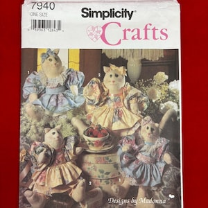 May include: A Simplicity Crafts 7940 sewing pattern envelope featuring four stuffed cat dolls wearing floral dresses with bows. The text includes Simplicity 7940, Crafts, and Designs by Madonna. The dolls are depicted in various outfits with lace and fabric details.