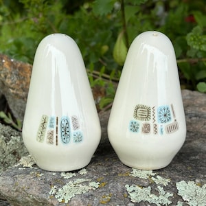 May include: A pair of off-white ceramic salt and pepper shakers with a rounded, tapered shape. Each shaker features a series of small, colorful graphic patterns in shades of light blue, brown, and green, including starbursts and geometric lines.