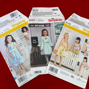 May include: Three Simplicity sewing pattern envelopes labeled 8063, 8522, and 8145. Each envelope features color photographs of children wearing dresses and carrying themed bags, including a blue bird, a snowman, and a lemon-shaped accessory.