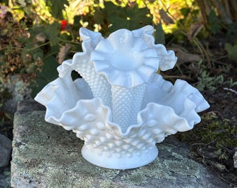 May include: A white milk glass epergne featuring a hobnail texture and ruffled edges. The centrepiece consists of a wide, bowl-shaped base with a flared, crimped rim and a central trumpet-shaped vase with a matching hobnail pattern and ruffled top.