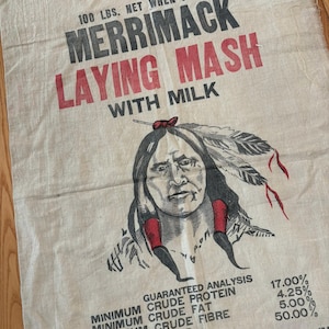 May include: A beige burlap sack featuring black and red text that reads Merrimack Laying Mash With Milk and 100 lbs. net when packed. A black and white illustration of a person with feathers in their hair is centered on the fabric.