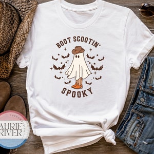 Peut inclure: T-shirt blanc avec un graphique d'un fantôme portant un chapeau de cowboy et des bottes. Le texte "BOOT SCOOTIN' SPOOKY" est imprimé sur le t-shirt.