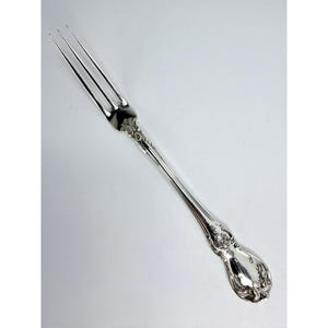 May include: A silver-colored metal fork featuring three long, slender tines and a handle decorated with intricate, embossed scrollwork and floral-inspired patterns. The surface of the utensil is polished to a high, reflective, and metallic sheen.
