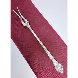 May include: A long, slender silver cocktail fork featuring two sharp tines and an ornate, embossed floral pattern on the handle. The metal surface is polished and reflective, showcasing intricate scrollwork details at the base of the tines and handle.