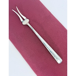 May include: A silver-toned metal cocktail fork featuring two slender, curved tines and a smooth, elongated handle. The utensil has a polished, reflective surface and a simple, streamlined design, showcasing a classic aesthetic for serving small appetizers.