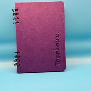 May include: A purple spiral-bound notebook with the word "Thankable." embossed vertically on the cover. The notebook has a textured surface and black metal spiral binding. The background is a solid light blue.
