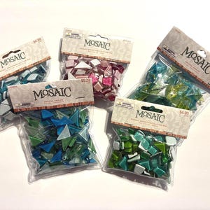 May include: Five clear plastic bags of mosaic tiles in various colors, including blue, green, pink, and white. Each bag has a cardboard header with the word "MOSAIC" printed on it. The tiles are small, irregular shapes.