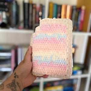 Może przedstawiać: Ręcznie robiony, prostokątny przedmiot na szydełku o miękkim, teksturowanym wyglądzie. Przedmiot posiada poziome paski w pastelowych kolorach: różowym, jasnoniebieskim i żółtym. Obramowanie jest w jednolitym, jasnobeżowym kolorze.