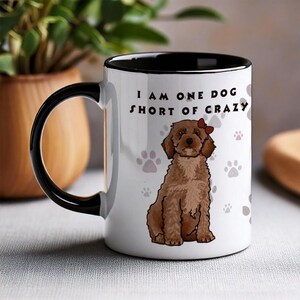 May include: A white ceramic mug with a black handle and interior rim. The mug features a brown curly-haired dog illustration with a small red bow on its head, surrounded by light grey paw prints and the text I AM ONE DOG SHORT OF CRAZY.