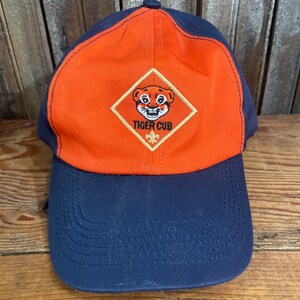 May include: A blue and orange baseball cap with a Tiger Cub logo embroidered on the front. The logo features a cartoon tiger with a yellow background.