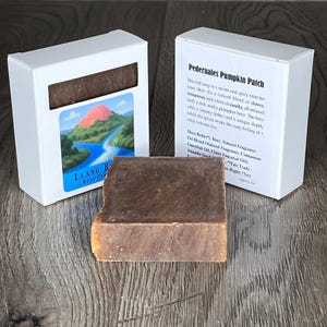 Puede incluir: Una barra de jabón rectangular marrón con un borde marrón claro, junto a una caja blanca con el texto "Pedernales Pumpkin Patch". La caja también tiene una imagen de paisaje y el texto "Llano River Soap Co."