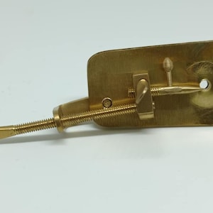 Könnte beinhalten: Ein messingfarbenes mechanisches Gerät mit rechteckiger Basis und Gewindestange. Es verfügt über einen Schraubmechanismus und einen kleinen Griff, was auf eine Verwendung zum Klemmen oder Einstellen hindeutet.