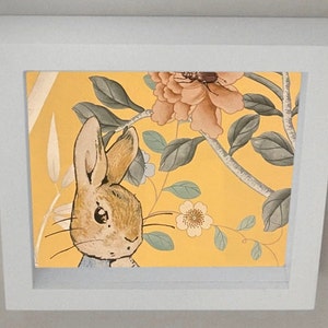 Peut inclure: Un cadre blanc avec un fond jaune représentant un lapin brun avec un ventre blanc et un motif floral dans les tons de rose, vert et blanc.