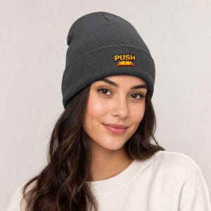 May include: A charcoal gray knit beanie featuring a cuffed brim. Centered on the front of the cuff is an embroidered graphic with the word PUSH in bold yellow letters above a stylized orange and red open book icon.