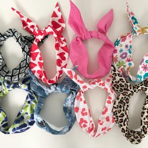 May include: Assortment of colourful fabric headbands with various patterns, including hearts, flowers, and animal print. The headbands are tied in a bow at the top.