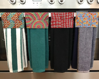 May include: Four hanging kitchen towels with patterned fabric tops and solid or striped absorbent bottoms. The towels feature green and white stripes, solid teal, solid black, and a textured grey fabric, each with a unique colourful printed top.