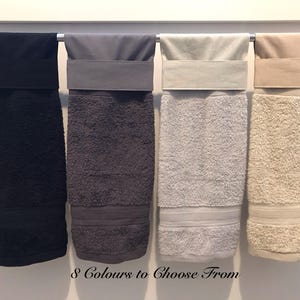 May include: Four hanging towels in black, dark grey, light grey, and beige. Each towel features a smooth fabric band at the top and a textured terry cloth lower section. The text "8 Colours to Choose From" is printed at the bottom of the image.