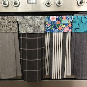 May include: Four decorative kitchen towels hanging on an oven. The towels display diverse patterns, including floral, striped, and geometric designs. The colours include grey, blue, white, and black. The towels are made of fabric and are designed for kitchen use.