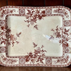 Könnte beinhalten: Rechteckige Servierplatte aus Keramik mit cremefarbenem Mittelteil und braunem Blumendesign. Das Tablett hat einen Rand mit abwechselnden Blumen- und geometrischen Mustern und Blumensträußen in jeder Ecke.