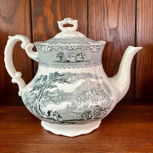 Puede incluir: Tetera de cerámica blanca con un diseño de paisaje en blanco y negro. La tetera presenta un asa curva, un pico y una tapa con un asa decorativa. El diseño del paisaje incluye árboles, un castillo y figuras.