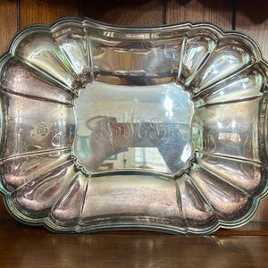 May include: A rectangular, silver-colored serving tray with a scalloped edge. The tray has a central, slightly recessed area and is engraved with initials. The tray is displayed on a dark wooden surface.