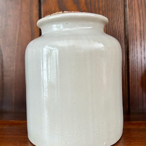 May include: A cream-colored, cylindrical ceramic jar with a cork stopper. The jar has a wide mouth and a slightly rounded shoulder. The surface is smooth and glossy, reflecting the light. The jar is sitting on a wooden surface.