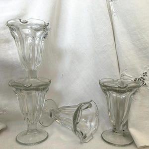 May include: Clear glass sundae dishes with a ruffled rim and a pedestal base. Three are upright, and one is lying on its side. The dishes are on a white background.
