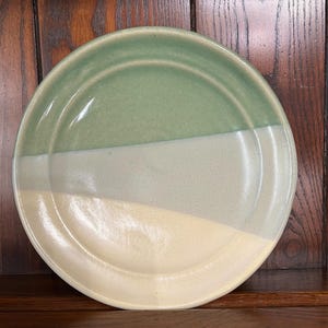 Może przedstawiać: Ręcznie robiony ceramiczny talerz z trójkolorowym wzorem. Górna część jest jasnozielona, ​​środkowa bladozielona, ​​a dolna kremowożółta. Talerz ma lekko podniesiony brzeg i gładkie, błyszczące wykończenie. Element dekoracyjny do kuchni.