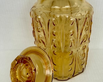 May include: Amber glass jar with a decorative embossed pattern and a matching lid. The jar has a wide mouth and a rounded body.