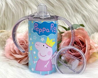 Pode incluir: Um copo de aço inoxidável azul e rosa com tampa de canudo e duas alças. O copo apresenta uma imagem de desenho animado de Peppa Pig vestida de fada com o texto "Peppa Pig" na frente.