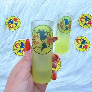 Puede incluir: Dos vasos de chupito transparentes y escarchados con un dise&ntilde;o de bal&oacute;n de f&uacute;tbol amarillo y rojo. El dise&ntilde;o presenta las letras "CA" y un mapa de las Am&eacute;ricas. Los vasos est&aacute;n sobre una superficie blanca y peluda con pegatinas amarillas y rojas con el mismo dise&ntilde;o.