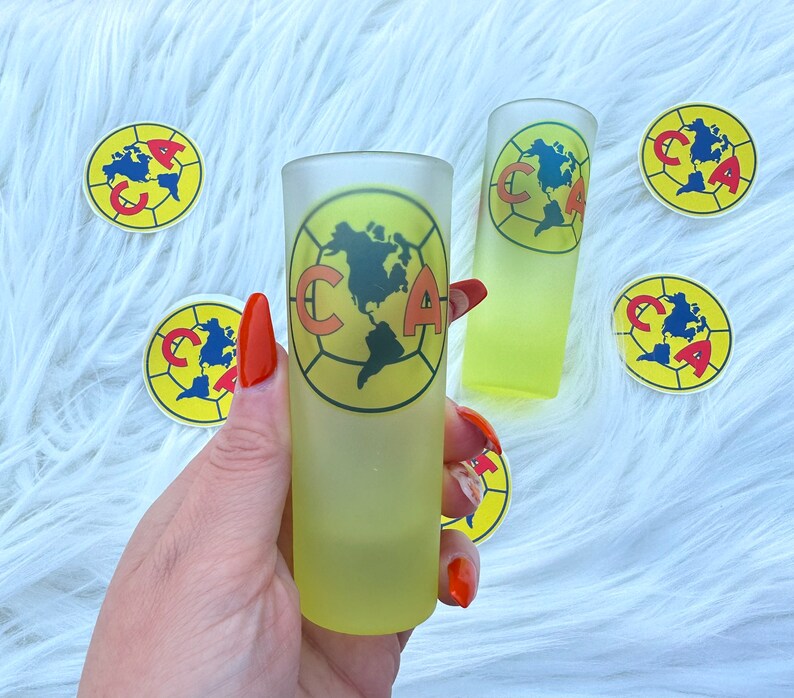 Puede incluir: Dos vasos de chupito transparentes y escarchados con un dise&ntilde;o de bal&oacute;n de f&uacute;tbol amarillo y rojo. El dise&ntilde;o presenta las letras "CA" y un mapa de las Am&eacute;ricas. Los vasos est&aacute;n sobre una superficie blanca y peluda con pegatinas amarillas y rojas con el mismo dise&ntilde;o.