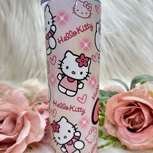 Puede incluir: Un vaso blanco con un dise&ntilde;o rosa de Hello Kitty. El vaso presenta m&uacute;ltiples im&aacute;genes de Hello Kitty con corazones rosas y brillos.