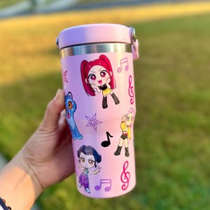 May include: A pink insulated tumbler with a silver rim and a pink lid. The tumbler is decorated with cartoon characters, musical notes, and stars. The tumbler is held in a hand.