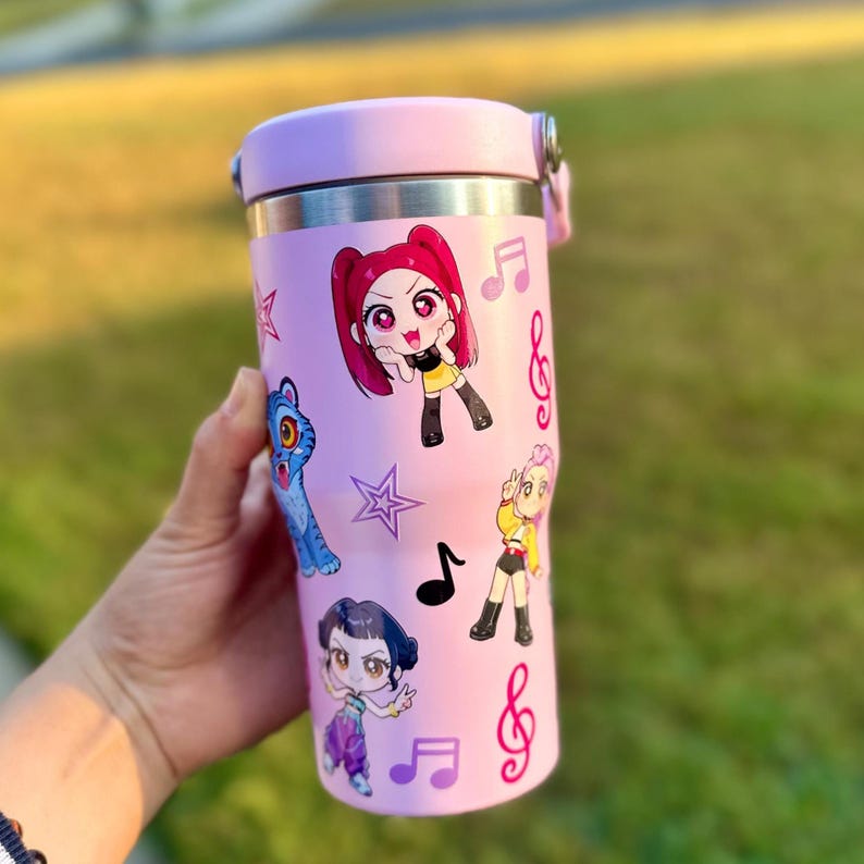 May include: A pink insulated tumbler with a silver rim and a pink lid. The tumbler is decorated with cartoon characters, musical notes, and stars. The tumbler is held in a hand.