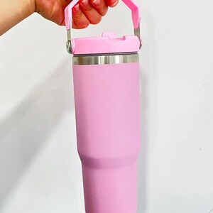 May include: A pink insulated tumbler with a matching handle and lid. The tumbler is held by a hand, showcasing its cylindrical shape and stainless steel accents. The handle is attached with silver metal hardware.