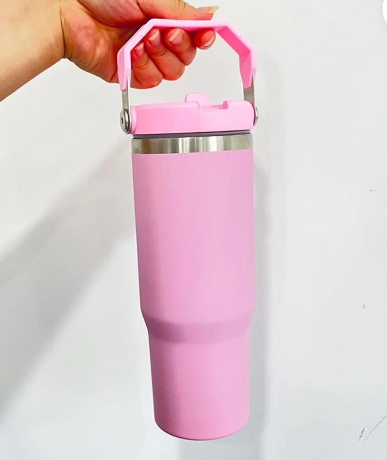 May include: A pink insulated tumbler with a matching handle and lid. The tumbler is held by a hand, showcasing its cylindrical shape and stainless steel accents. The handle is attached with silver metal hardware.