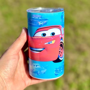 May include: A red and blue stainless steel tumbler with a Cars character, Lightning McQueen, on the front. The tumbler has a blue background with other Cars characters in the background.
