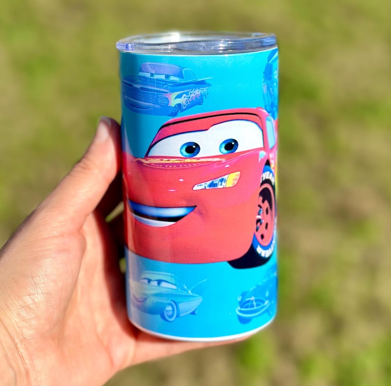 May include: A red and blue stainless steel tumbler with a Cars character, Lightning McQueen, on the front. The tumbler has a blue background with other Cars characters in the background.