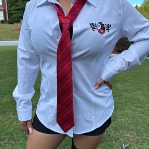 May include: White button-down shirt with a red and navy striped tie. The shirt has a small emblem on the chest. The tie is knotted at the neck. The shirt is worn with black shorts and black boots.