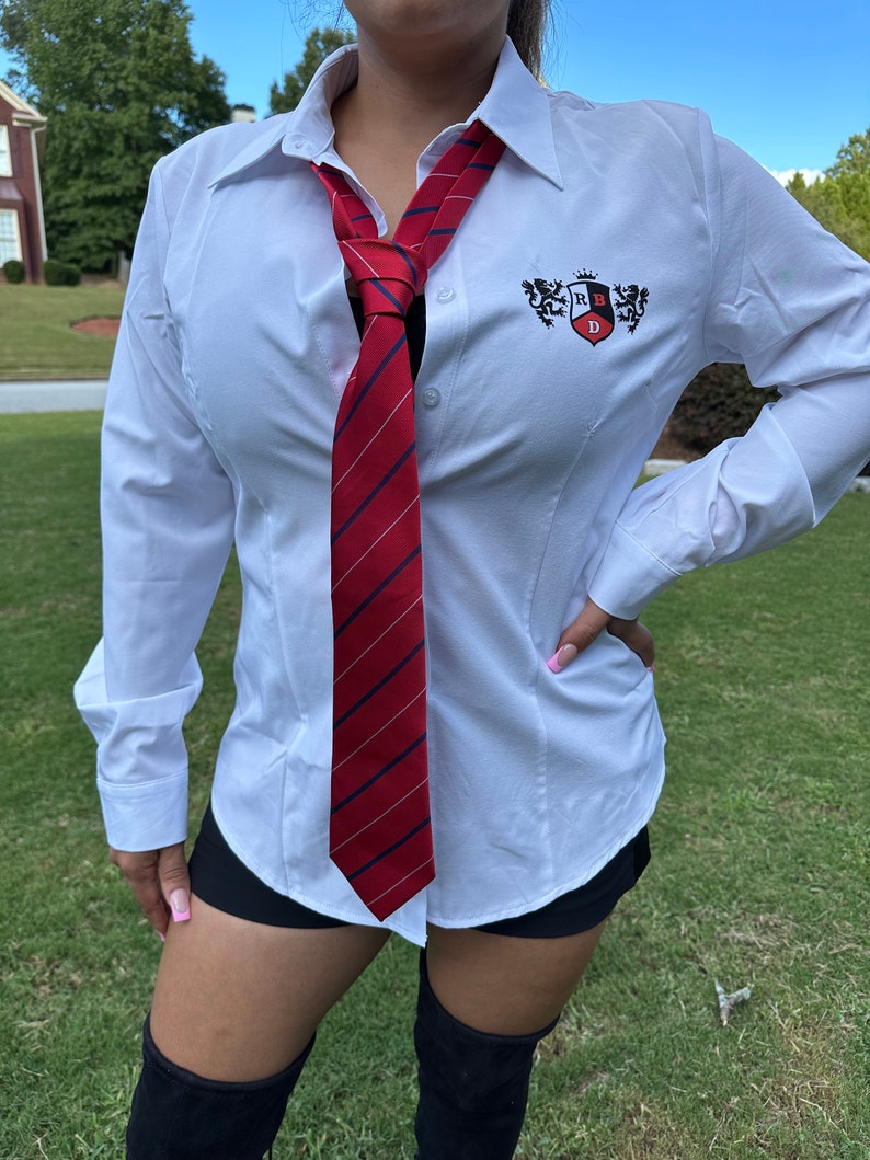 May include: White button-down shirt with a red and navy striped tie. The shirt has a small emblem on the chest. The tie is knotted at the neck. The shirt is worn with black shorts and black boots.