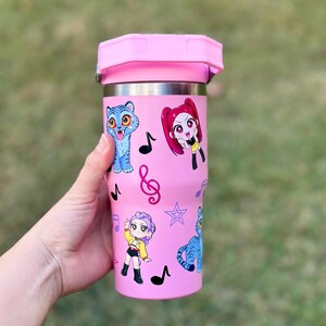 May include: Pink insulated tumbler with a pink lid, featuring cartoon illustrations of characters and musical notes. The tumbler is held by a hand, showcasing the design. The tumbler is approximately 7 inches tall.