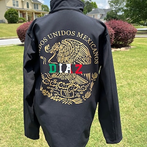 Pode incluir: Casaco preto de softshell com um brasão mexicano bordado a ouro que apresenta uma águia, um cacto e uma cobra. O texto "DOS UNIDOS MEXICANOS" está escrito em círculo em torno da águia. O texto "DIAZ" está escrito em vermelho, branco e verde abaixo da águia.