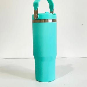 May include: A teal insulated tumbler with a handle and a screw-on lid. The tumbler has a stainless steel rim and a matching teal handle. The tumbler is designed to keep beverages cold or hot. The tumbler is approximately 9 inches tall.