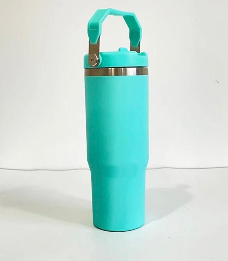 May include: A teal insulated tumbler with a handle and a screw-on lid. The tumbler has a stainless steel rim and a matching teal handle. The tumbler is designed to keep beverages cold or hot. The tumbler is approximately 9 inches tall.