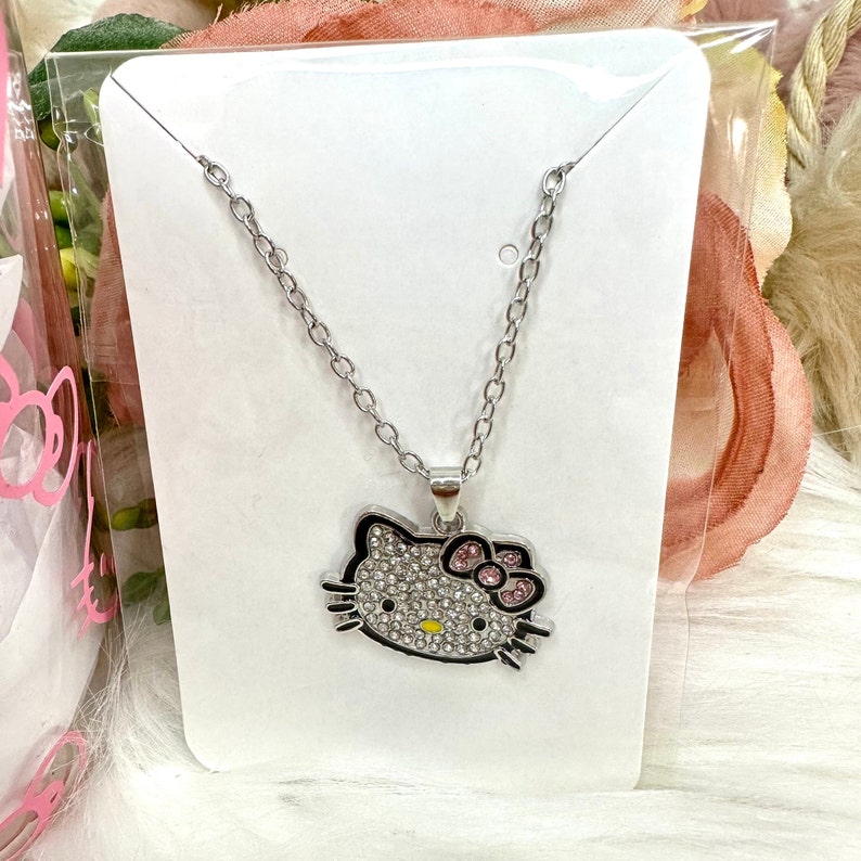 Puede incluir: Un collar de plata con un dije de Hello Kitty. El dije est&aacute; cubierto de piedras de cristal transparentes y tiene un lazo rosa.