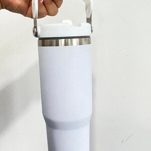 May include: A white insulated tumbler with a white handle and lid. The tumbler has a stainless steel band near the top. The handle is attached to the lid with silver metal hinges.