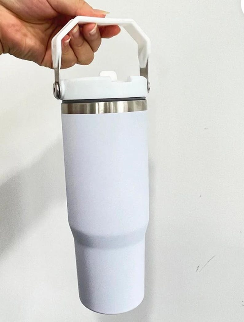 May include: A white insulated tumbler with a white handle and lid. The tumbler has a stainless steel band near the top. The handle is attached to the lid with silver metal hinges.