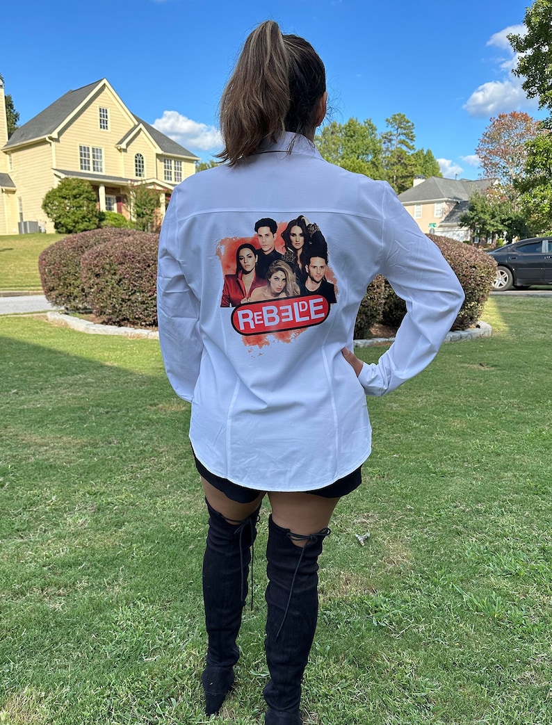 May include: White button-down shirt with a colorful graphic of a group of people and the word "REBELDE" in red and white. The shirt is long-sleeved.
